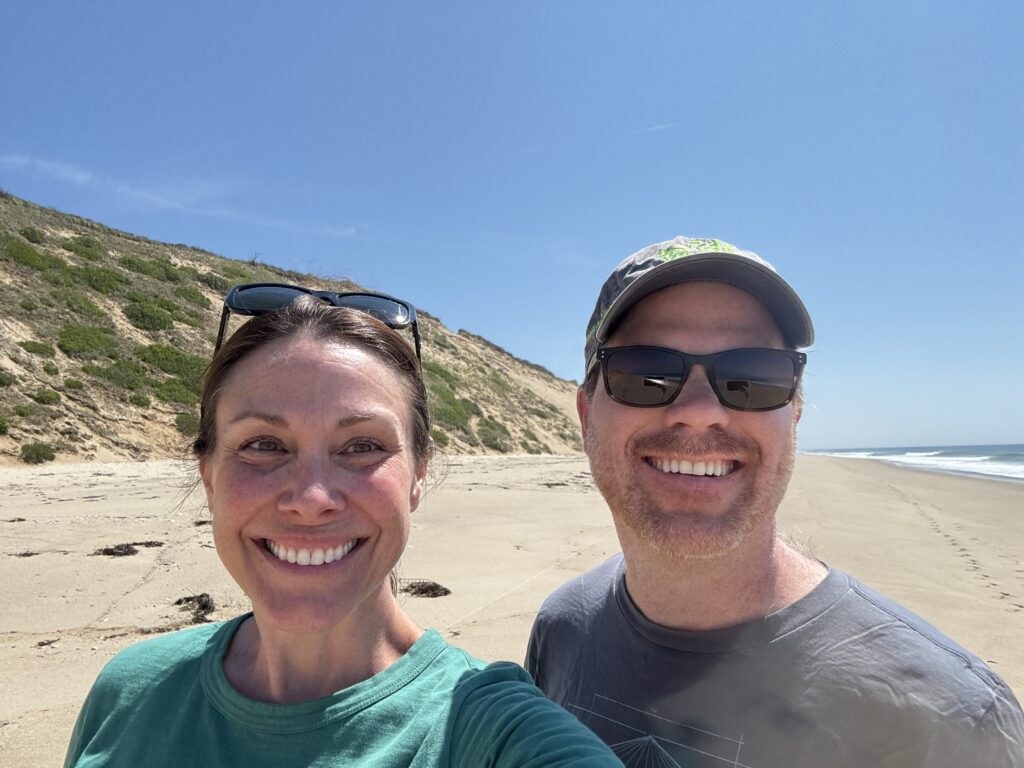 Jesse Brackenbury (President and CEO, Statue of Liberty & Ellis Island Foundation) and Julia Bateman (Executive Director, Friends of Cape Cod National Seashore) stand on a sandy beach on a sunny day. You can see gentle waves crashing on shore behind them.